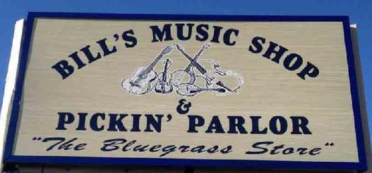 Exterior sign that reads Bill's Music Shop & Pickin' Parlor, The Bluegrass Store. The sign is white, with a dark blue border and lettering, and with a dark blue graphic of a guitar, fiddle, banjo, upright bass, and mandolin.