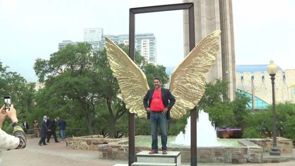 A man in a red shirt poses with giant bronze wings displayed in an outdoor art exhibition