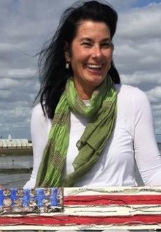 Deane Bowers, dressed in a white top with green scarf, holds a piece of art resembling the U.S. flag.