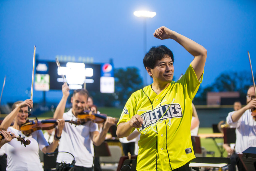 Nakahara, wearing a neon yellow Columbia Fireflies jersey, conducts the orchestra at the Fireflies' ballpark at dusk.
