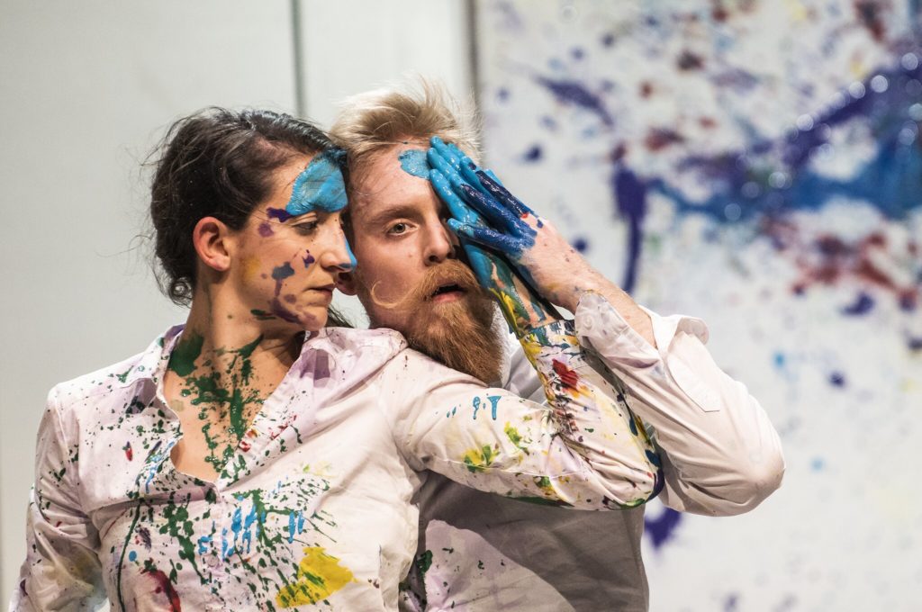 A woman and man, splattered in bright paint, stand close to perform a theatre production.