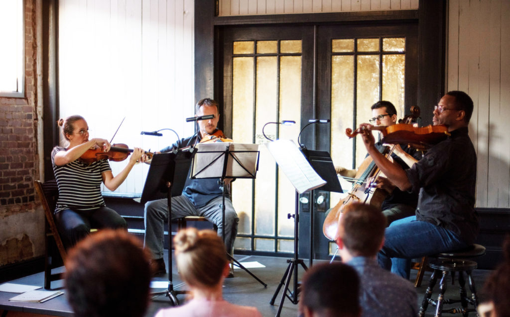 A string quartet plays music in a rustic indoor setting in front of a small seated group.