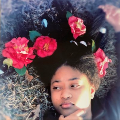 Photograph of a young Black girl lying in the grass with hibiscus blooms in her hair.