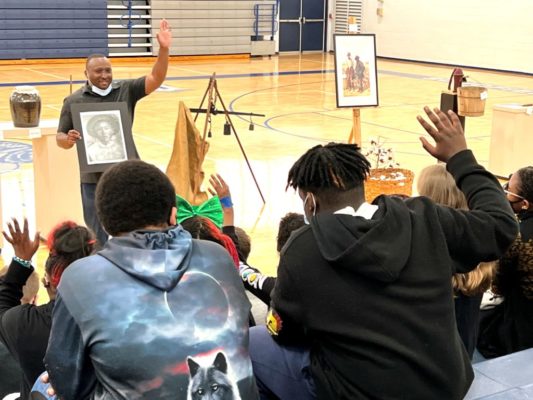 Dr. Curry speaks to students from the floor of the school gymnasium as they look on from bleachers. 