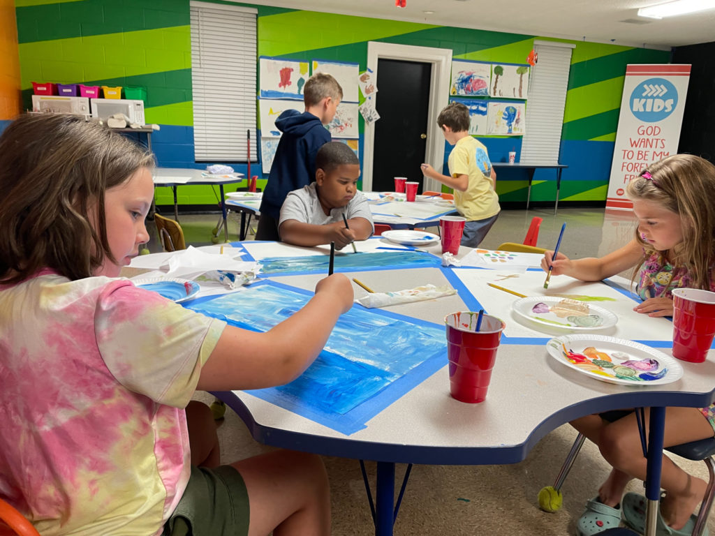 Children seated at a low table painting landscapes in a classroom.