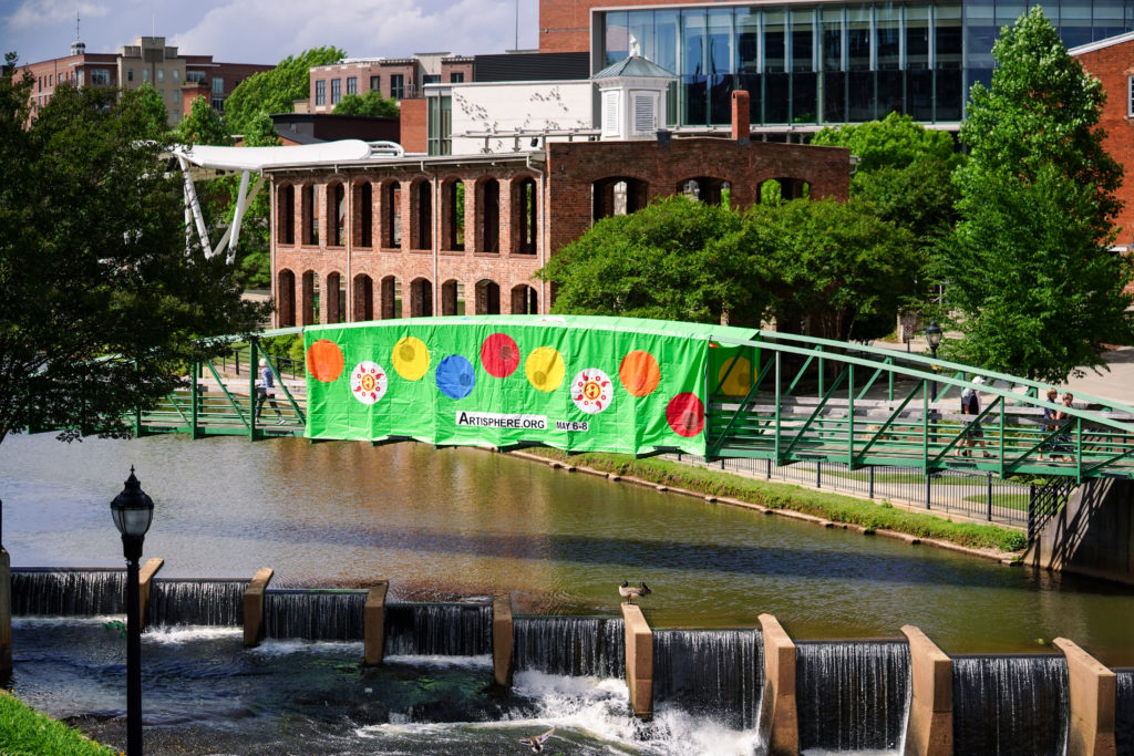 A green banner with colorful dots drapes over a footbridge over the Reedy River in Greenville.
