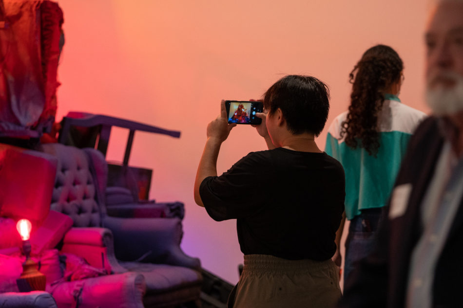 In a dimly-lit room, an art patron with short black hair in a black t-shirt snaps a mobile phone picture of South Carolina State Fellow Brittany M. Watkins' "Maladaptation Sits," on exhibit at the launch of the tour.