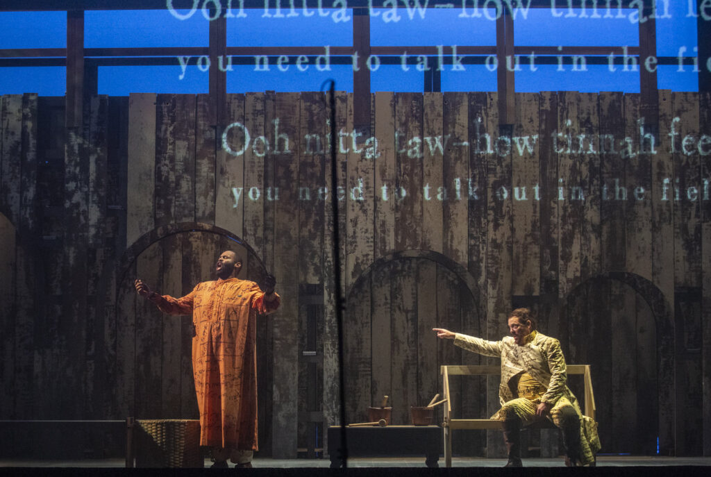A Black man with close-cropped hair and a beard, wearing a flowing orange tunic, sings on the left of a darkened stage. A white man with short dark hair and white flowing clothes points at the Black man while singing. The stage set looks like a weathered wooden-slat fence with text from the opera in white superimposed via projection.