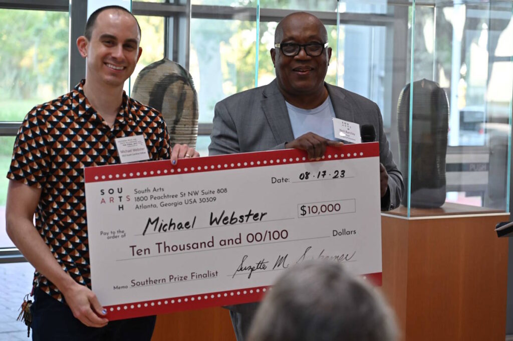 Two men stand side by side, holding a prop presentation check to South Carolina artist Michael Webster, holding the check on the left side, at an indoor presentation in front of large windows.