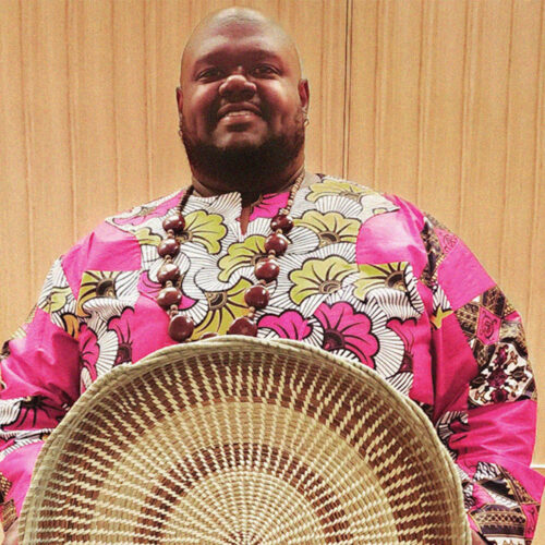 A man, smiling proudly and wearing a brightly-colored patterned shirt, holds an intricately-woven sweetgrass basket.