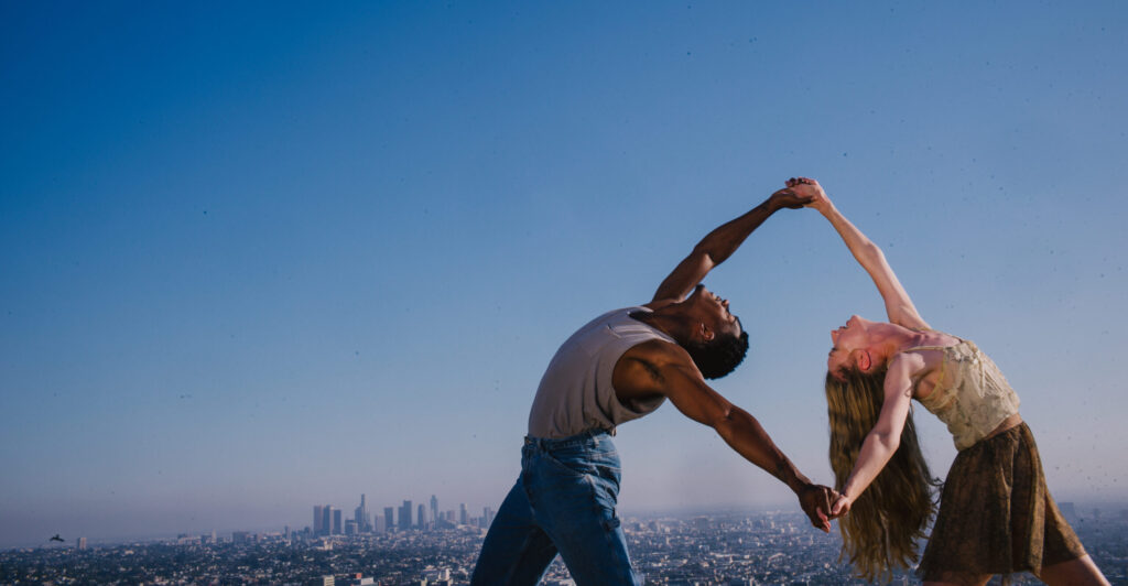 Two dancers dance to Romeo in Juliet, outdoors against a bright blue sky with the Los Angeles skyline far in the background