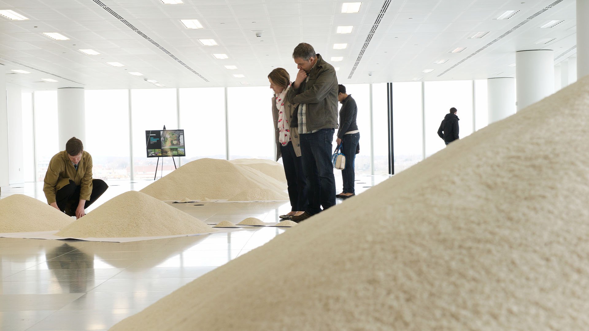 People look at large mounds of uncooked rice, displayed in a white, light-filled room.