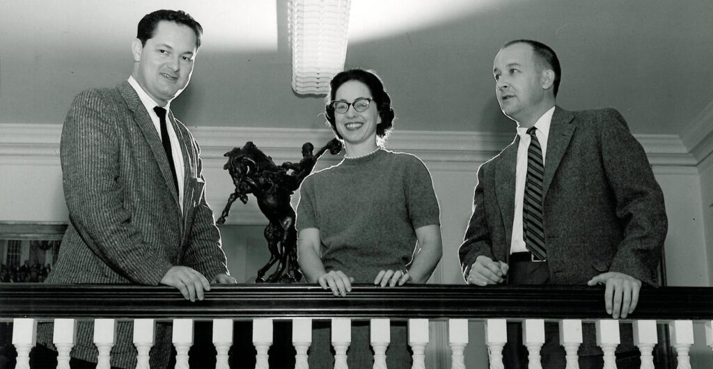 Three White people stand indoors, behind a wooden railing, facing the camera in this black and white image.