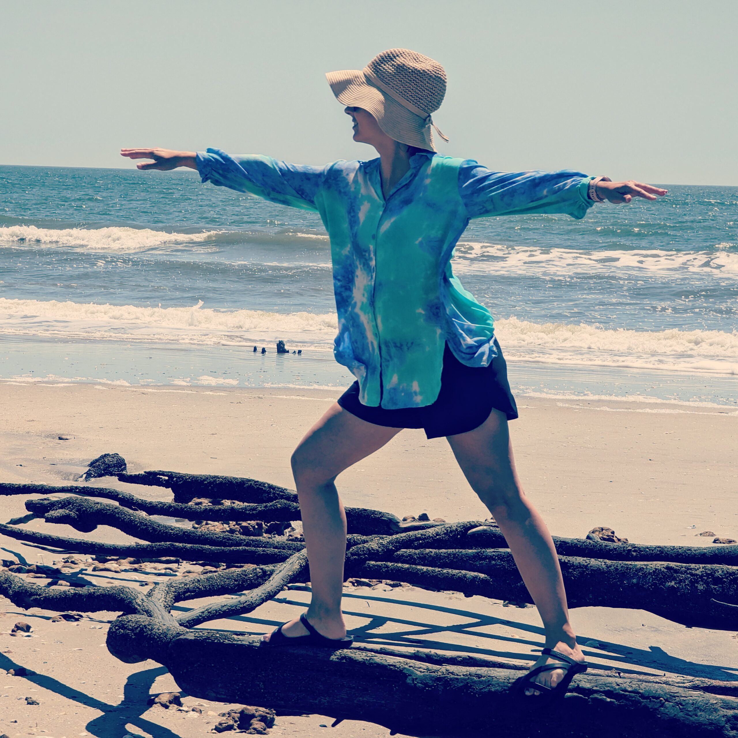 a woman in casual clothing holds a yoga pose among driftwood on a beach
