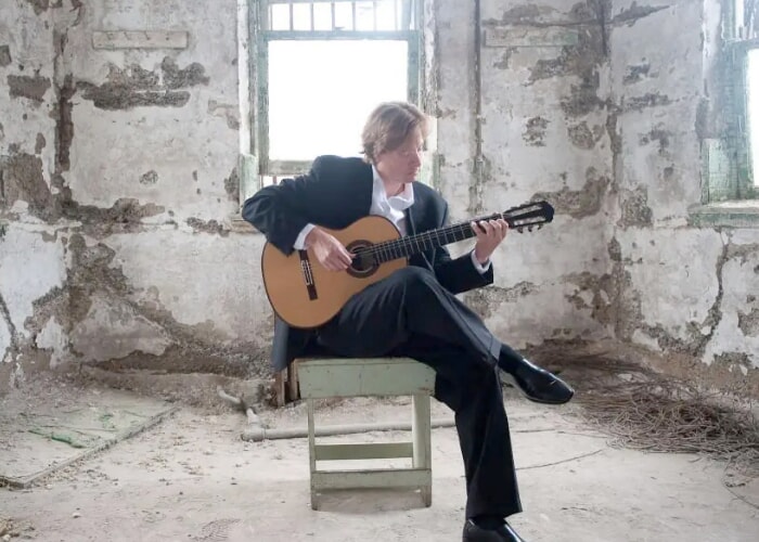 a seated blond individual plays acoustic guitar in a room with peeling plaster and windows