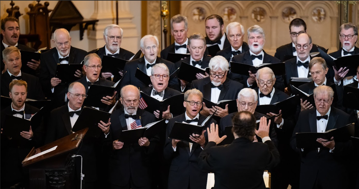 24 men in formal dress sing with black music folders, conducted by another man, near a church altar