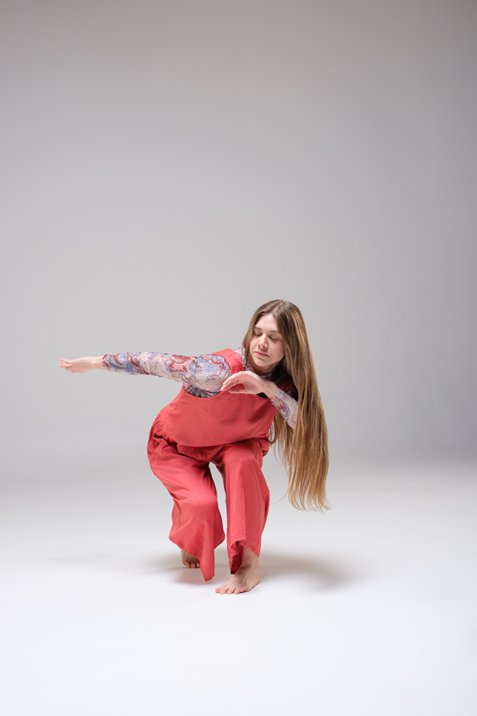 A dancer with long blonde hair crouches in an angular pose. She wears pink overalls, a patterned shirt, and no shoes.