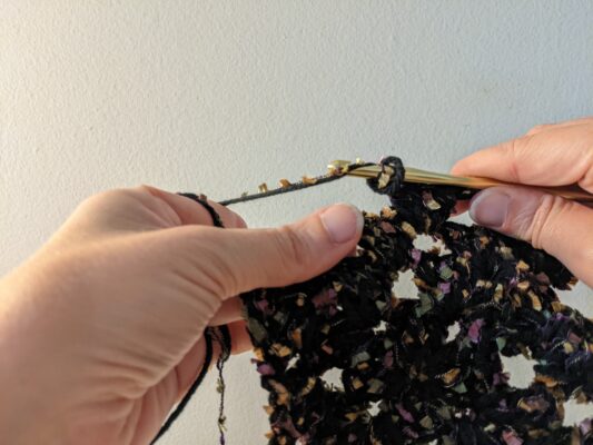 Hands use a gold crochet needle with dark yarn flecked with jewel tones.