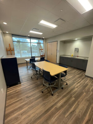 The studio at North Charleston Cultural Arts Department includes a large double window with medium dark hardwood flooring, two perpendicular tables with rolling chairs, and an inset credenza.