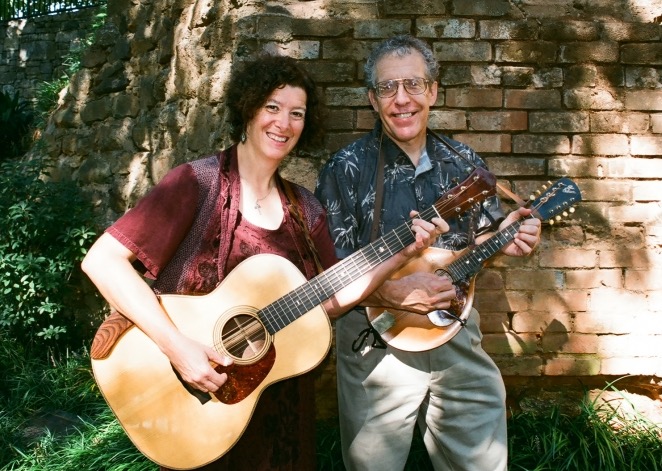 a man and woman standing in front of a brick wall holding guitar and a mandolin