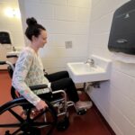 A white woman with dark hair pulled up in a bun in a black wheelchair attempting to use the sink in the old accessible restroom.
