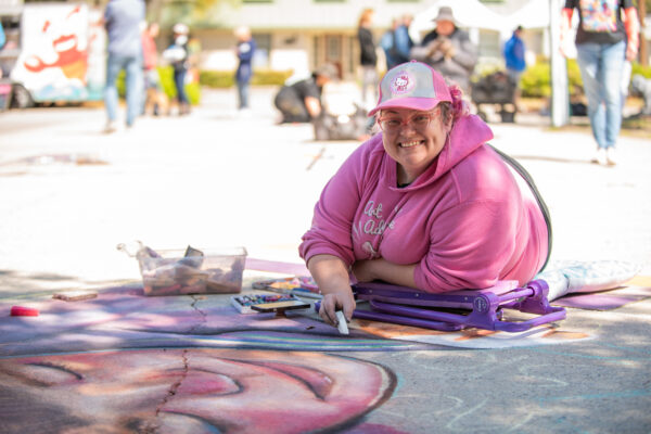 A smiling person lies on the pavement to make a complex chalk drawing of a face.