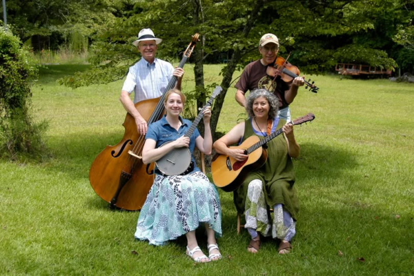 A 4-person string band poses outdoors with an upright bass, banjo, acoustic guitar, and fiddle.