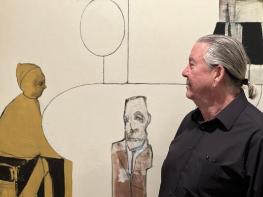 A man with a short gray ponytail looks at a work of art on a wall.