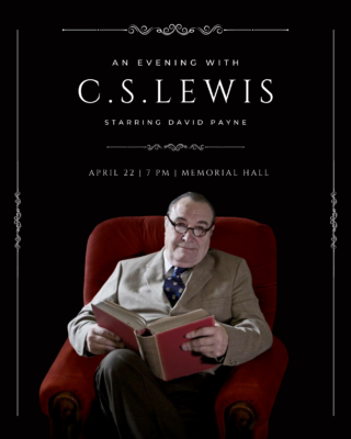 A man portraying author C.S. Lewis sits in a chair with a large hardcover book open on his lap.