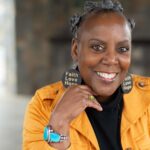 A Black woman in a yellow jacket smiles at the camera. She wears earrings that read Faith Love Hope.