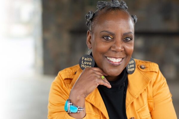 A Black woman in a yellow jacket smiles at the camera. She wears earrings that read Faith Love Hope.