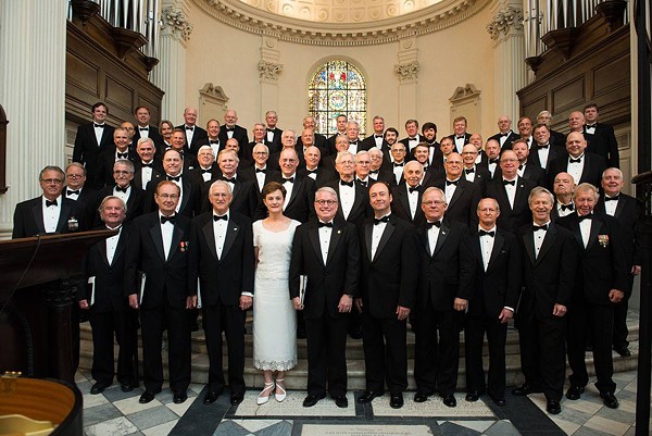 A large men's choir in formal dress posing in a church venue.