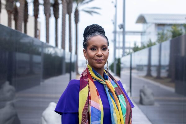 A Black woman wearing bright colors smiles confidently, standing outdoors.