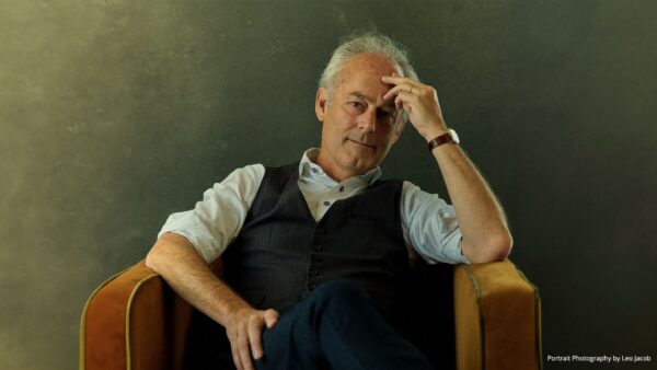 A man with gray hair sits in a chair with one hand resting on his forehead and a contemplative expression.
