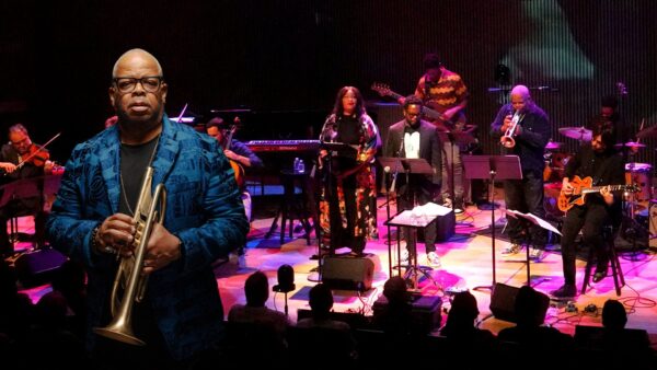 A man presenting as Black, holding a trumpet. In the background is an onstage band in front of an audience.