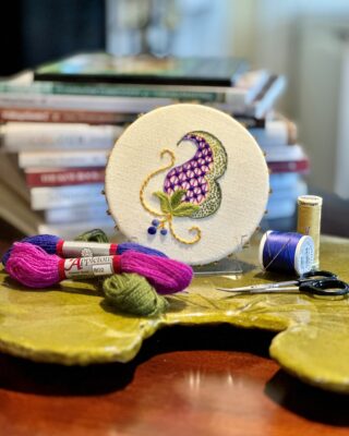 Fabric in a small hoop is embroidered with a curved shape in purple, blue, green, and yellow.
