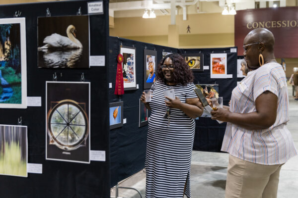 Two Black women enjoy an art show, looking at works on black fabric standing panels.