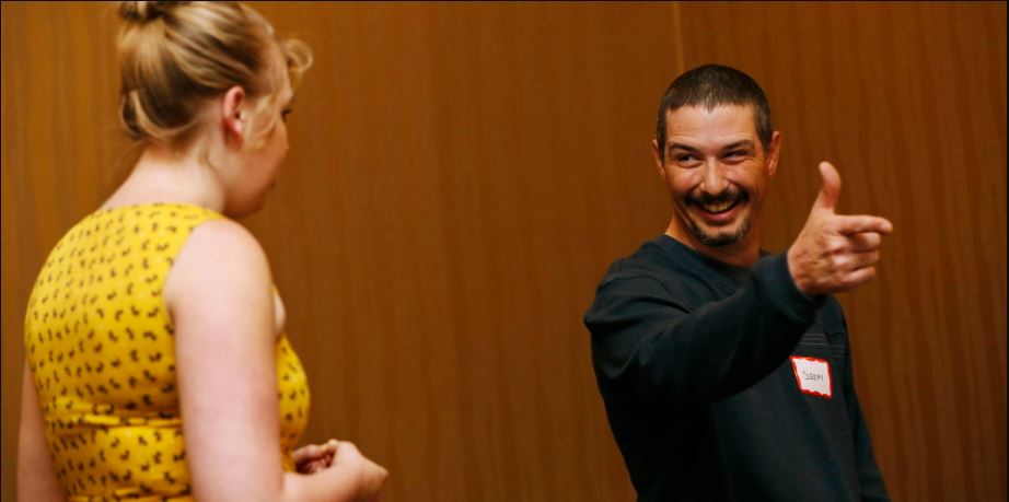 A man is smiling and miming a gun with one hand, as a woman looks on.
