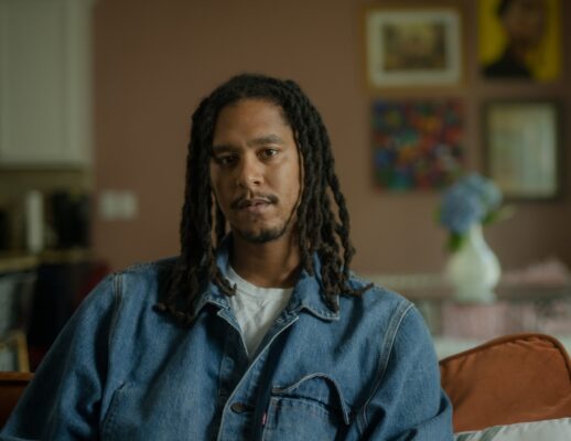 A Black man with dreadlocks looks at the viewer. He is wearing a denim shirt.