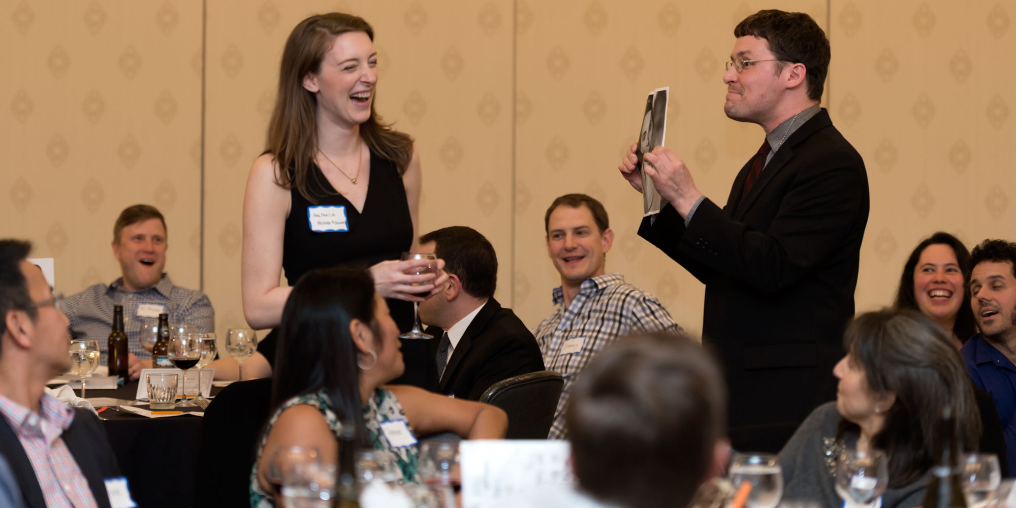 A woman in a dress laughs at a photo held by a man in a suit, amid dining tables.