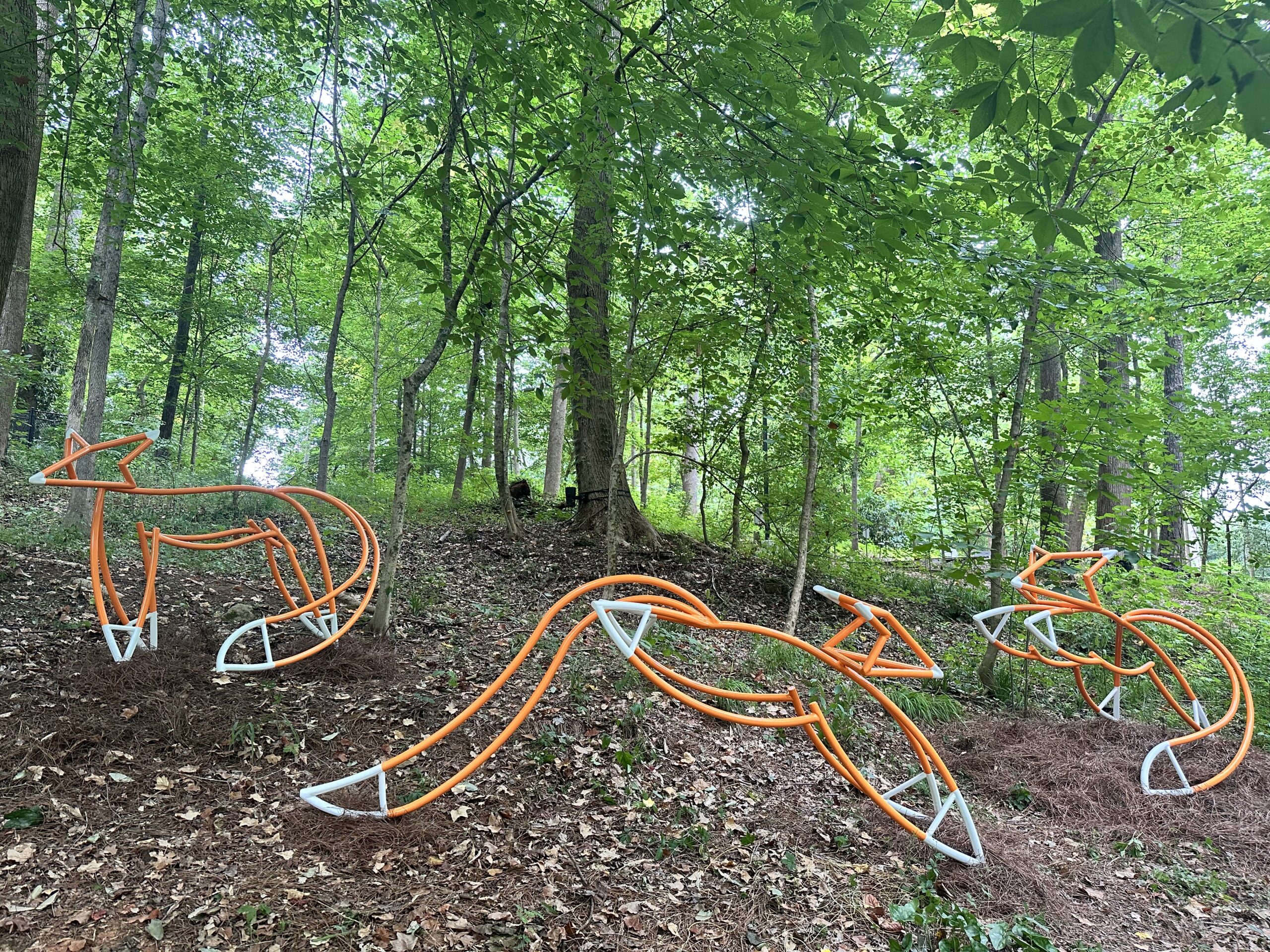Orange and white metal sculptures of a fox family in the forest.
