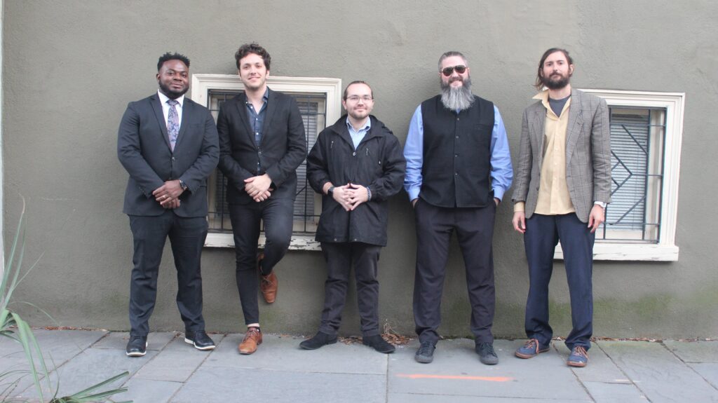 5 smiling men stand against an outdoor wall with windows.