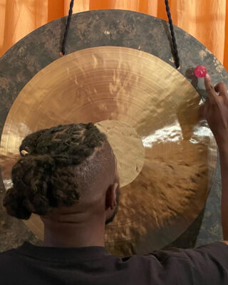 A Black person with short braided hair plays a large gong with a small pink mallet.