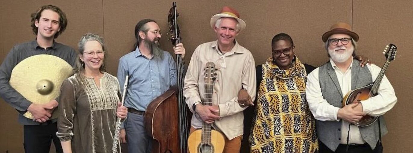 Smiling musicians stand in a row with a cymbal, flute, upright bass, acoustic guitar, and mandolin.