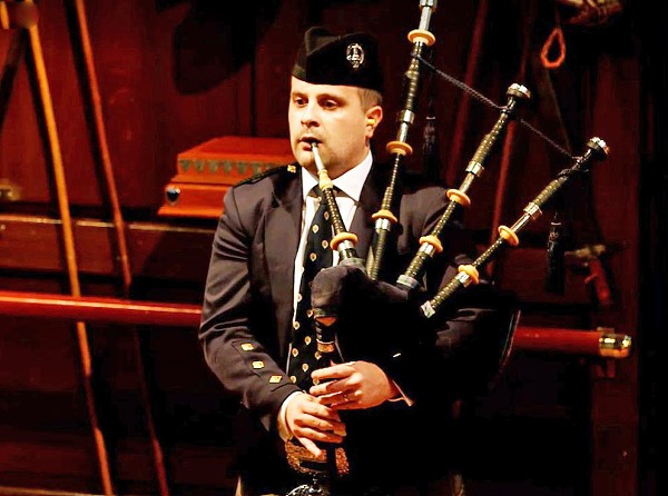 A man in full regalia plays bagpipes in a formal interior space.