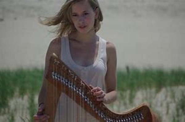 A blonde woman with a standing harp on a windy beach.