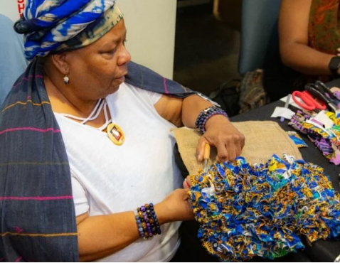 A seated Black woman in a headwrap works on a rag rug in tones of blue and yellow.