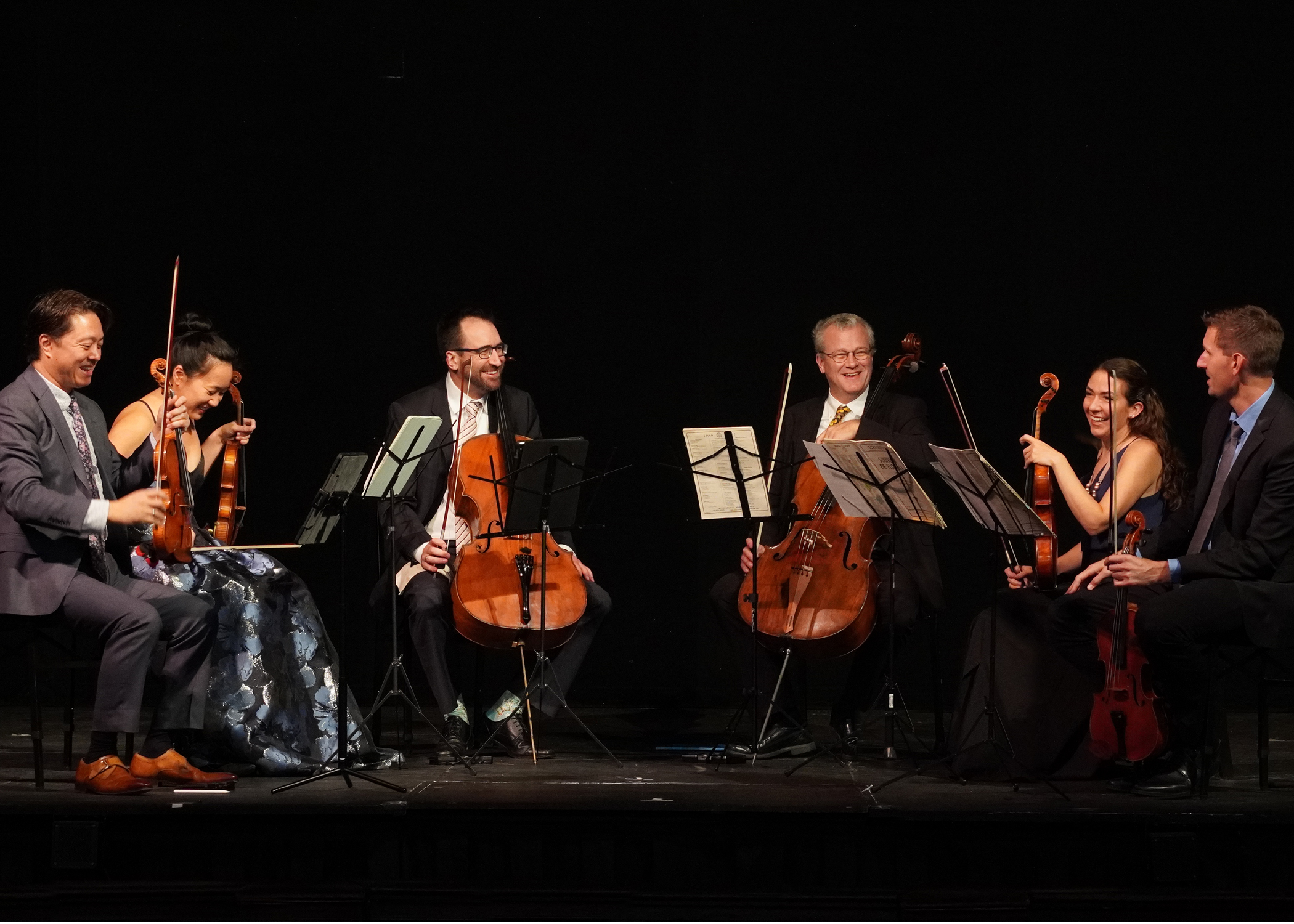 Members of a string sextet sit on stage with their instruments, smiling and laughing.