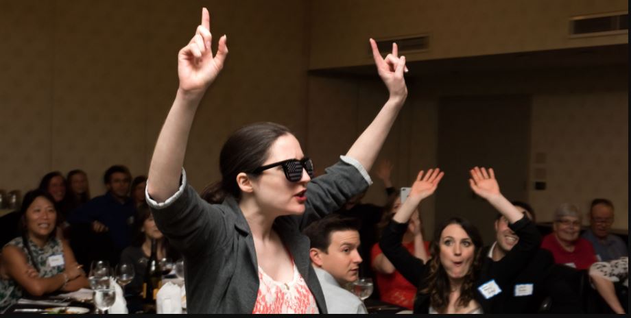 A woman in sunglasses and a blazer has her hands in the air at a dinner theatre performance.