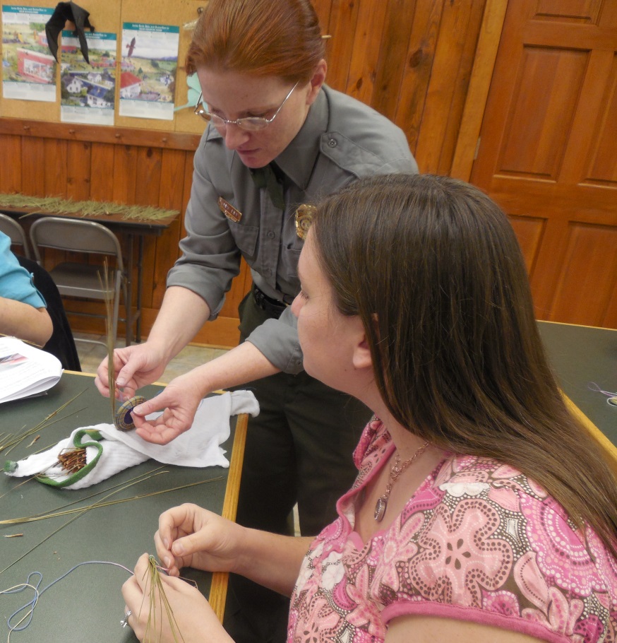 A uniformed person demonstrates coiling pine needles as another individual looks on.